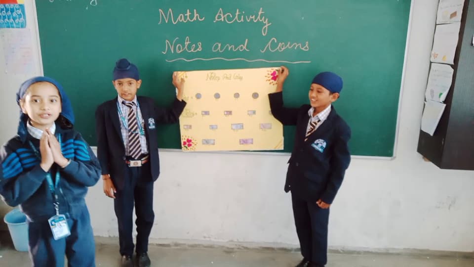Three young students presenting a notes-and-coins maths chart at the blackboard